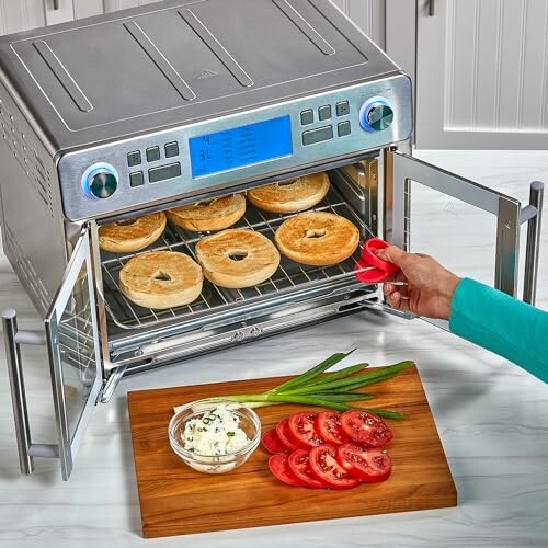 Countertop oven with bagels and sliced vegetables on a board.