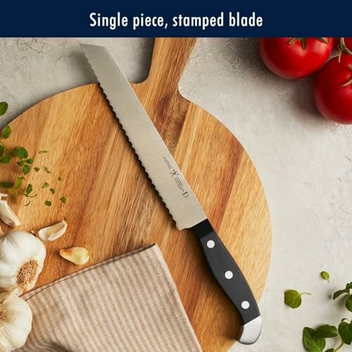 A serrated knife on a round wooden cutting board with tomatoes, garlic, and herbs.